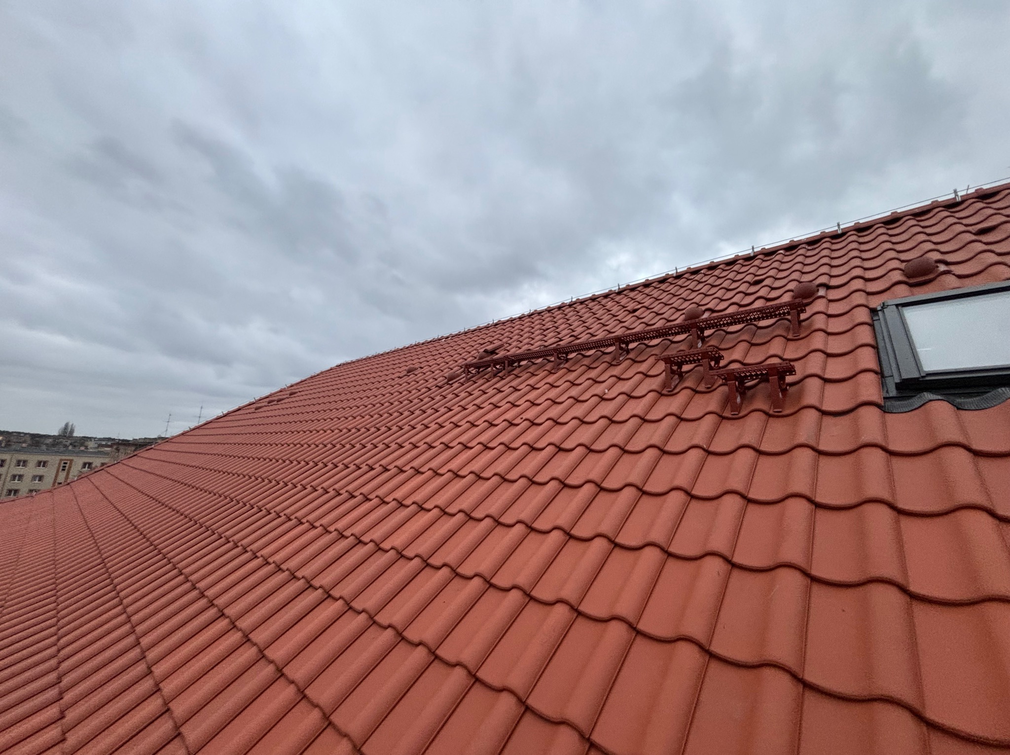 Czerwona dachówka ceramiczna z zamontowanymi stopniami kominiarskimi i oknem dachowym na tle pochmurnego nieba. Widok z dołu.