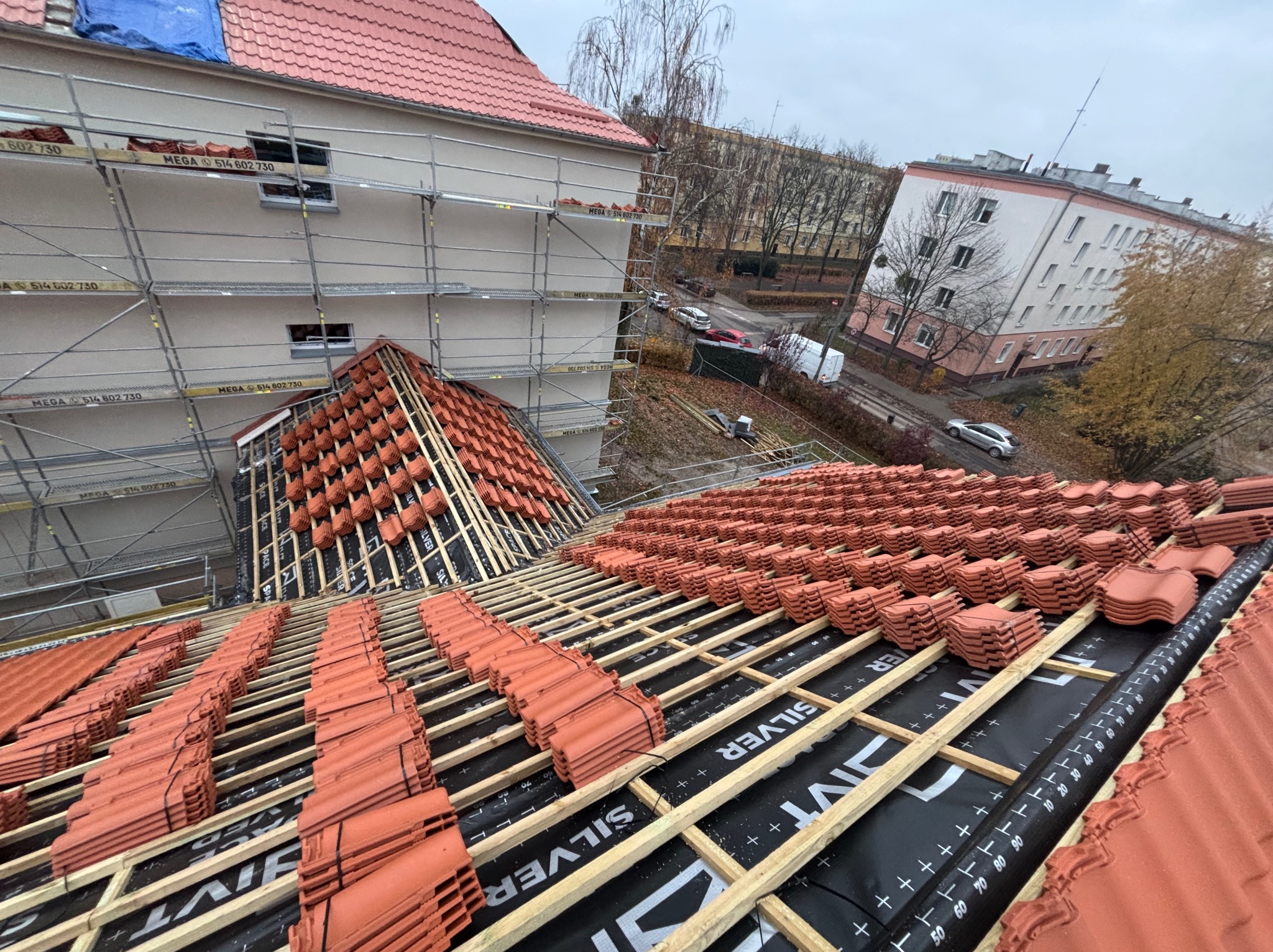 Montaż czerwonej dachówki na łatach z membraną Silver. Widok z góry na budynek w rusztowaniach. Częściowo pokryty dach. Jesienna pogoda.