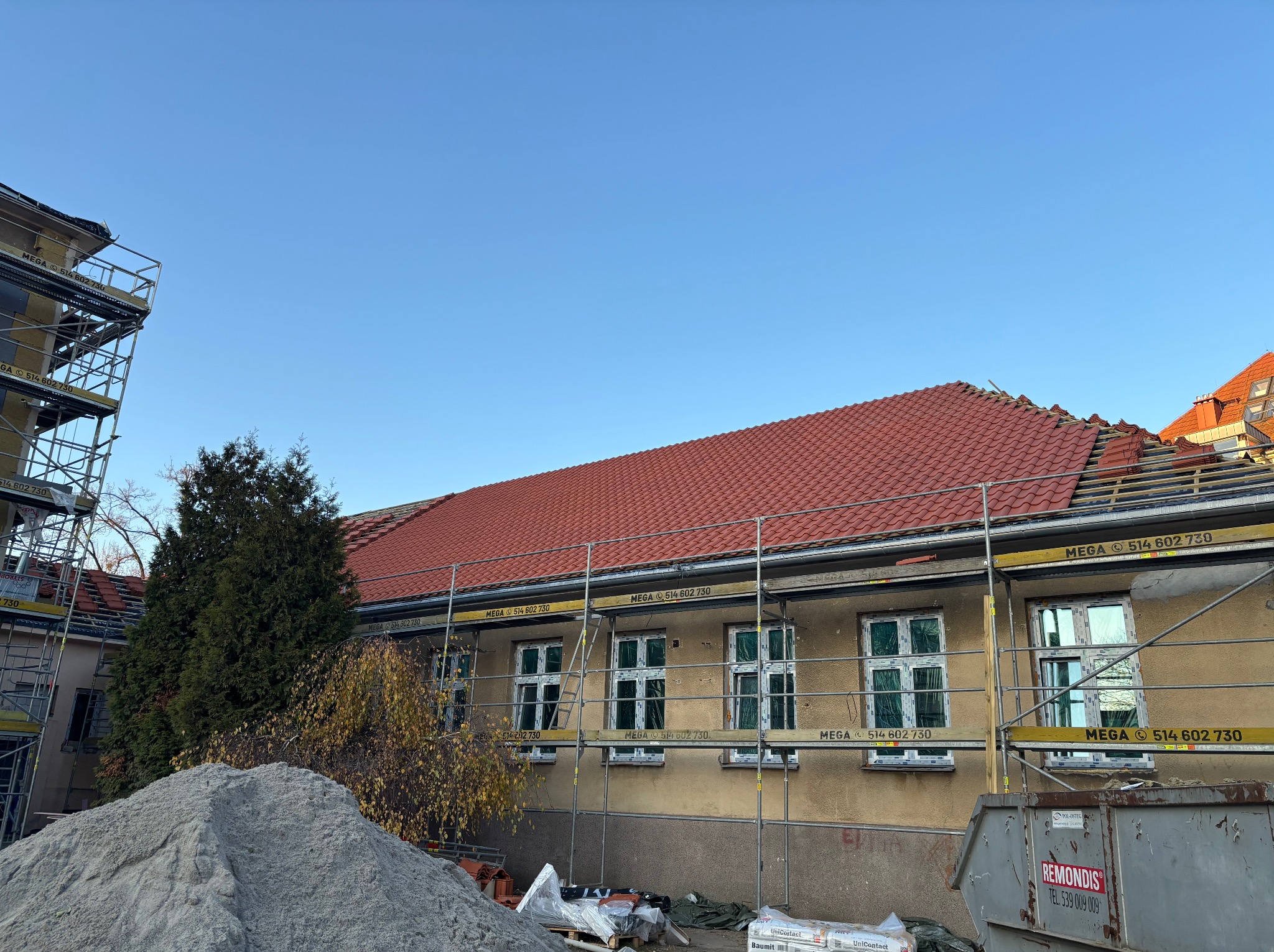 Budynek w trakcie remontu z rusztowaniami, nową czerwoną dachówką i oknami zabezpieczonymi folią. Na pierwszym planie hałda piasku i kontener na gruz.