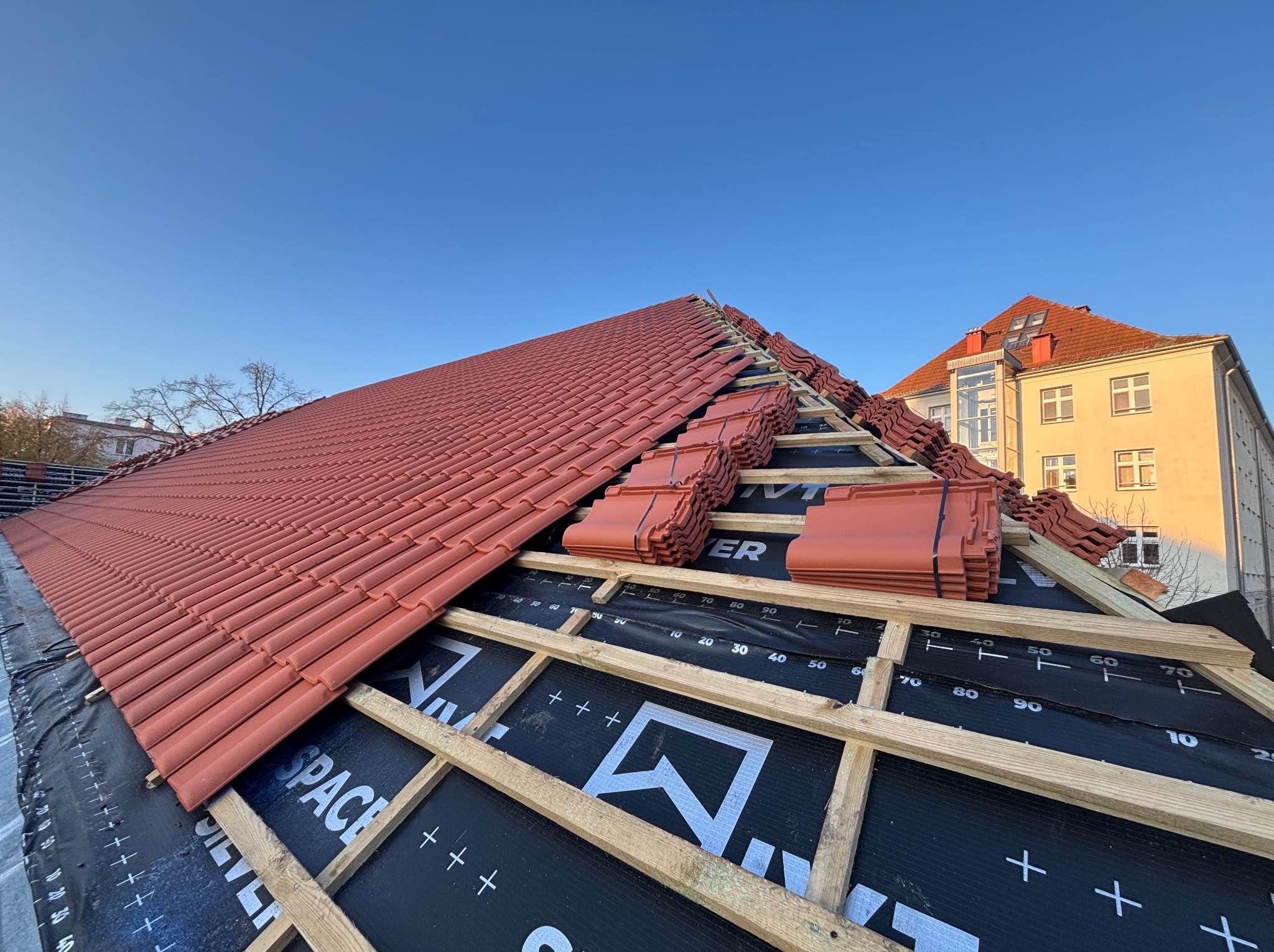 Częściowo pokryty dach z czerwoną dachówką, widoczne drewniane łaty i czarna membrana dachowa z nadrukiem, budynek w tle, błękitne niebo.