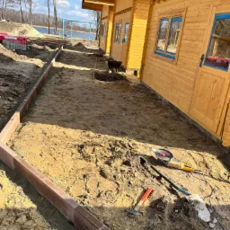 Przygotowanie podłoża pod ścieżkę z obrzeżami wzdłuż drewnianego domu, widoczne narzędzia: szlifierka kątowa, młotek, łopata. W tle jezioro i plac zabaw.