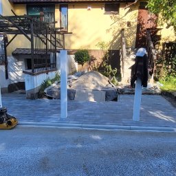 Usługi Brukarsko-Ziemne Oleh Berezovskyi - Częściowo ułożona szara kostka brukowa z żółtą zagęszczarką i białymi słupkami w ogrodzie przed domem. Widoczny piasek i ziemia, etap prac.