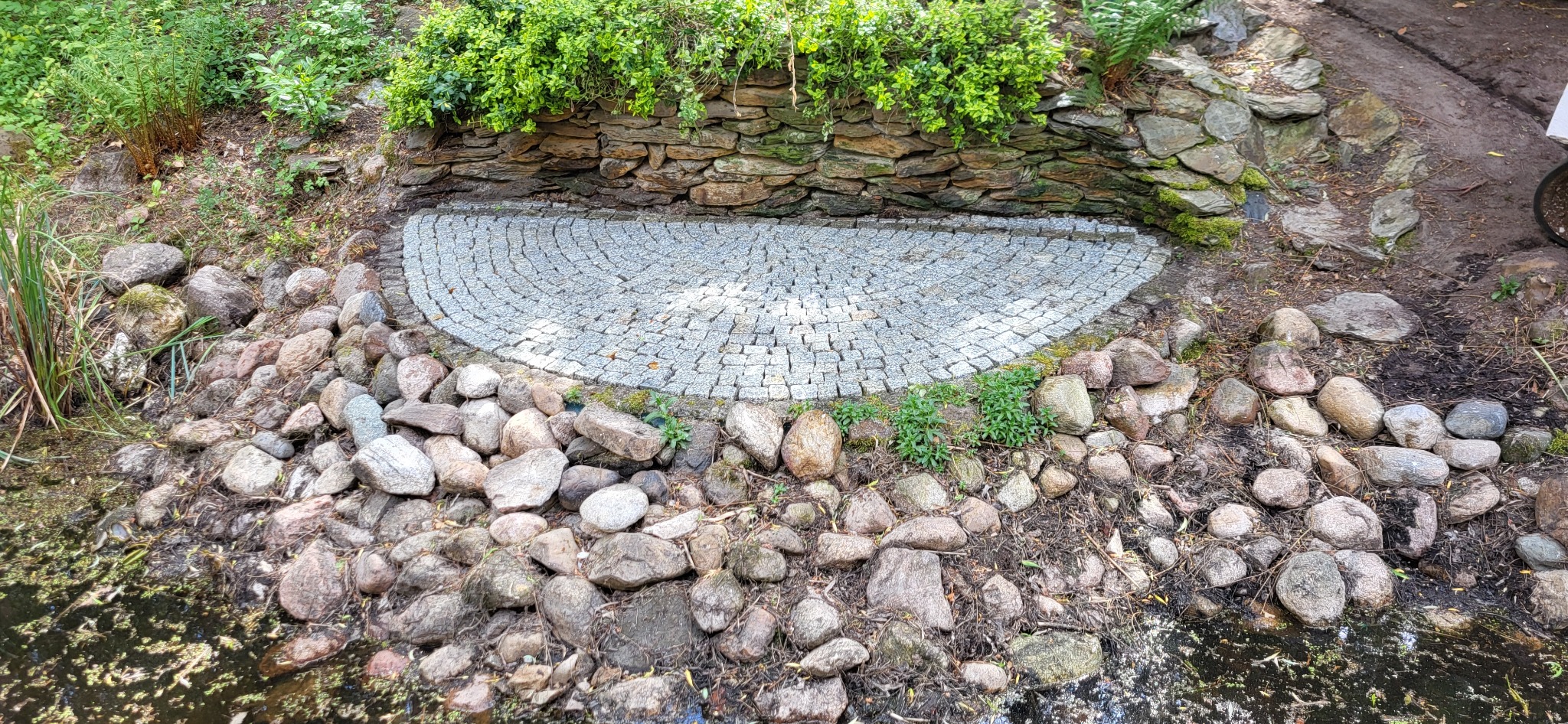 Półokrągła powierzchnia wyłożona drobną, szarą kostką granitową, otoczona większymi kamieniami i roślinnością w ogrodzie.
