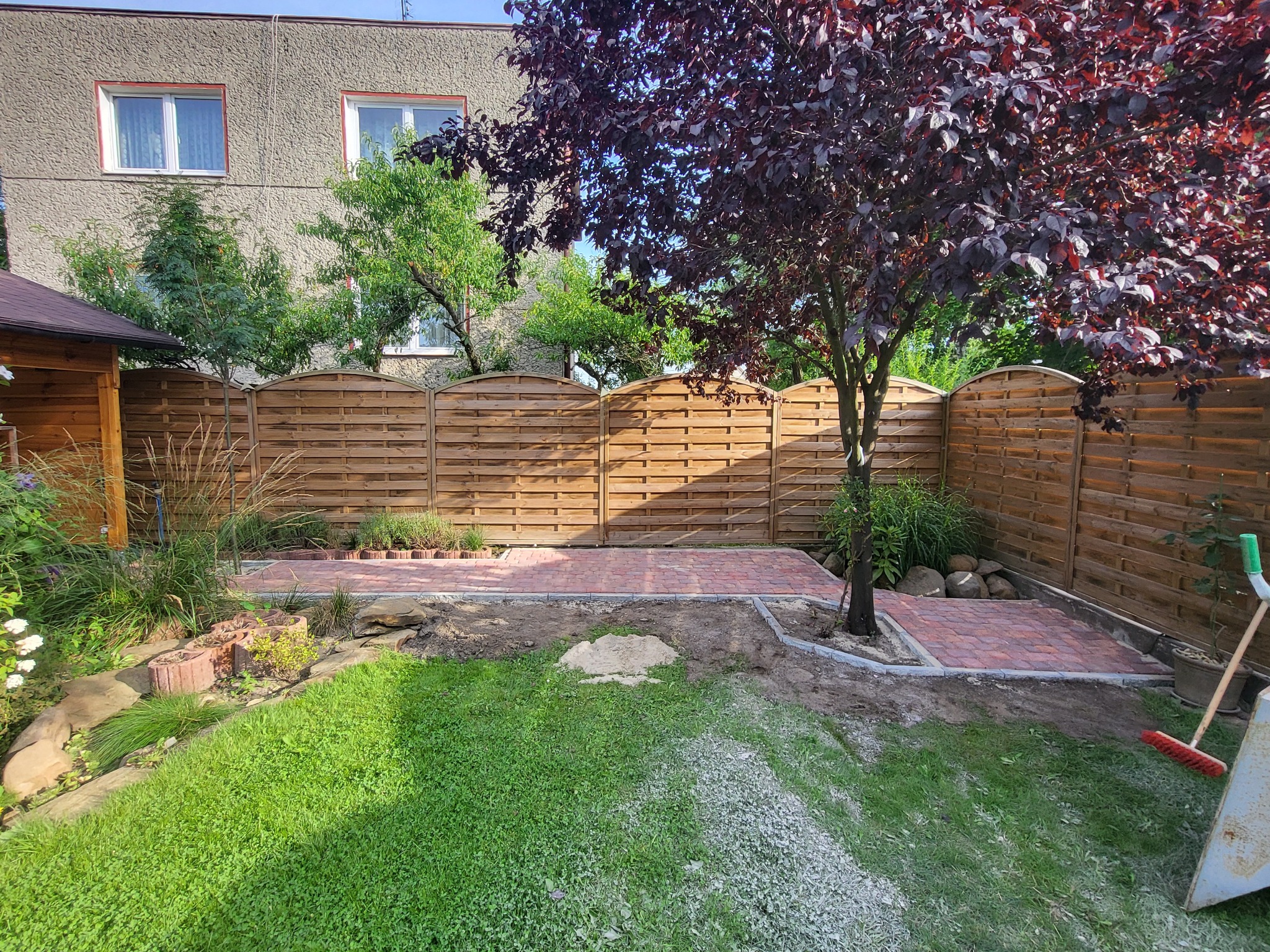 Fragment ogrodu z fragmentem ścieżki z czerwonej kostki brukowej w trakcie budowy, widoczny piasek, obrzeża i narzędzia ogrodnicze, w tle drewniany płot i drzewo o purpurowych liściach.