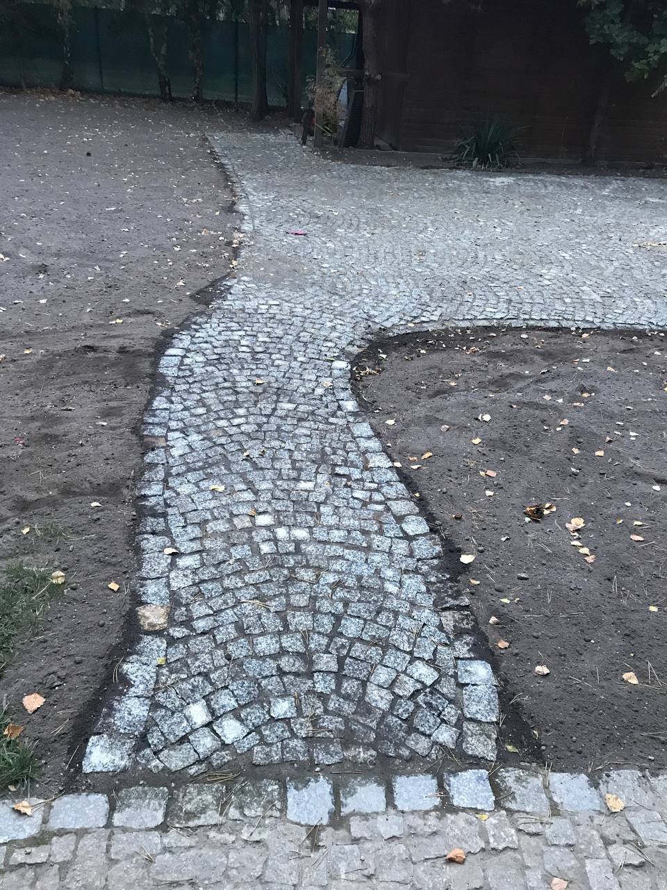 Ścieżka z małej kostki granitowej, układana łukiem na ziemi z opadłymi liśćmi, widok z góry, częściowo ukończona.