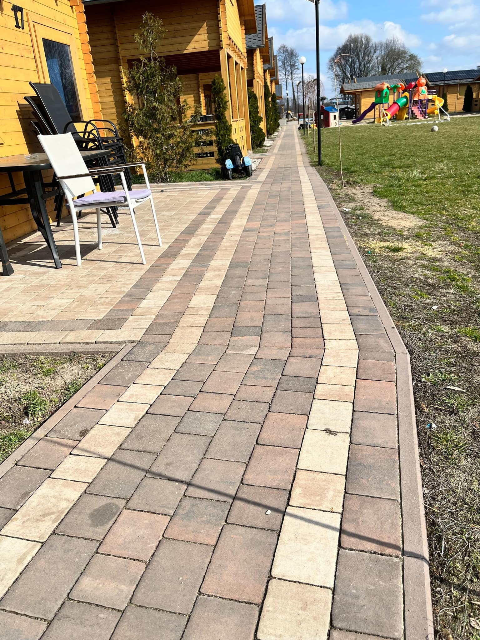 Długa, wijąca się ścieżka z kostki brukowej w odcieniach beżu i brązu prowadząca wzdłuż żółtych domków letniskowych w kierunku placu zabaw.
