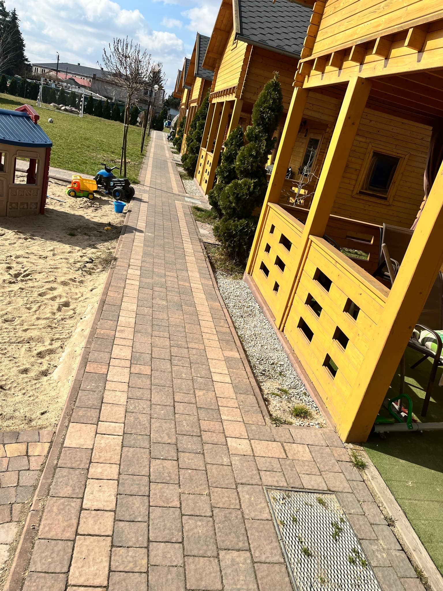 Długa, brązowo-szara ścieżka z kostki brukowej prowadząca wzdłuż rzędu żółtych, drewnianych domków letniskowych z małymi balkonami, obok plac zabaw z piaskiem i zabawkami.
