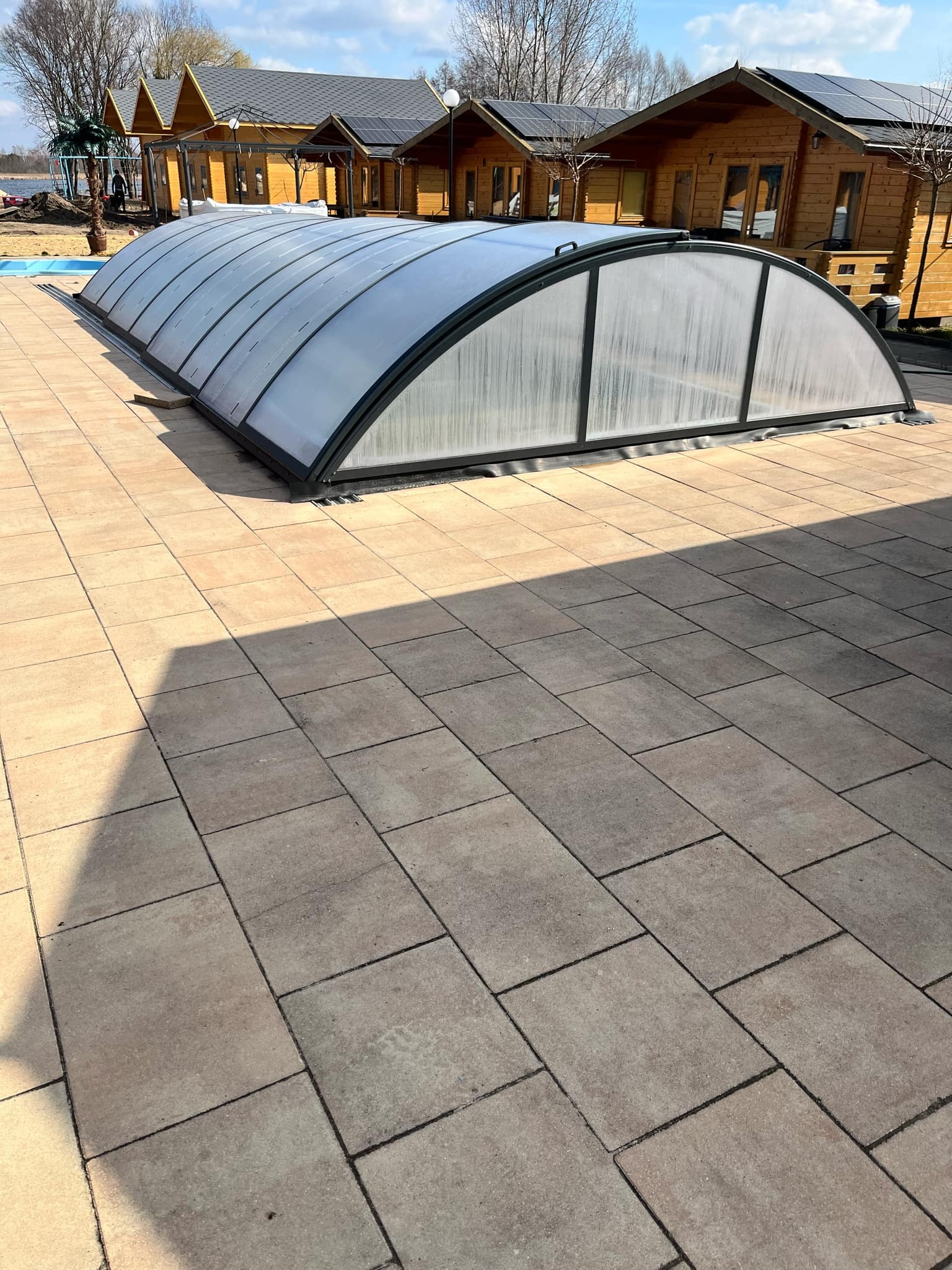 Jasnobrązowa kostka brukowa ułożona na dużej powierzchni z zadaszeniem basenowym w tle, w oddali widoczne drewniane domki.