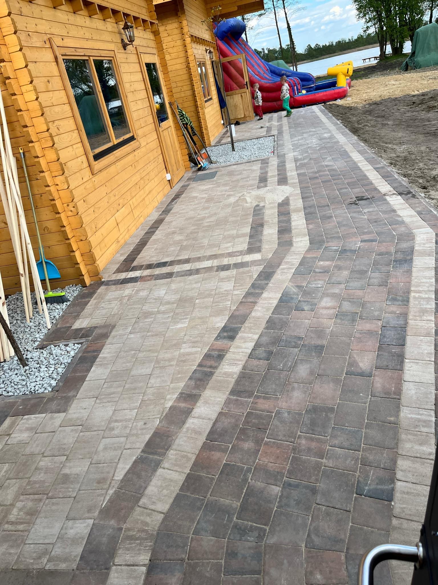 Ułożona kostka brukowa w geometryczny wzór przed drewnianym domkiem, z widocznym fragmentem dmuchanej zjeżdżalni w tle i dwójką dzieci.