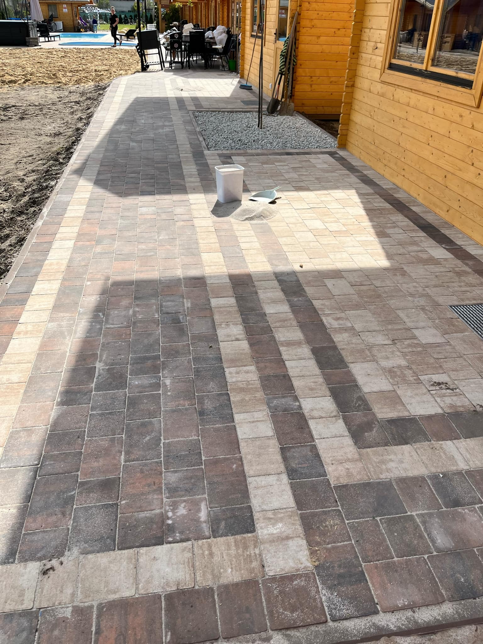 Ułożona kostka brukowa w odcieniach brązu i beżu, tworząca wzór na ścieżce obok drewnianego budynku, z widocznym białym wiadrem i szufelką.