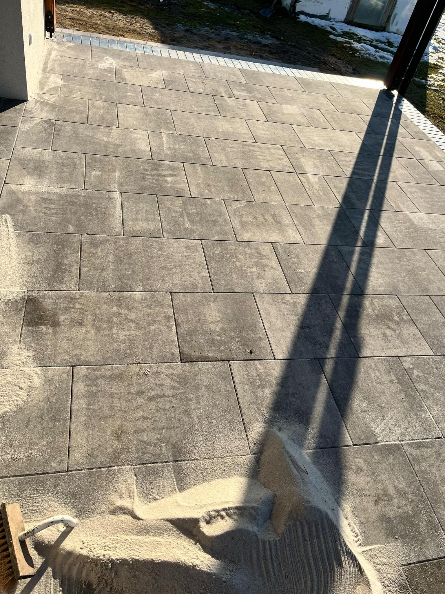 Świeżo ułożona, szara kostka brukowa z piaskiem rozsypanym na powierzchni, przygotowana do spoinowania. Widoczny cień słupka oraz fragment szczotki do zamiatania.
