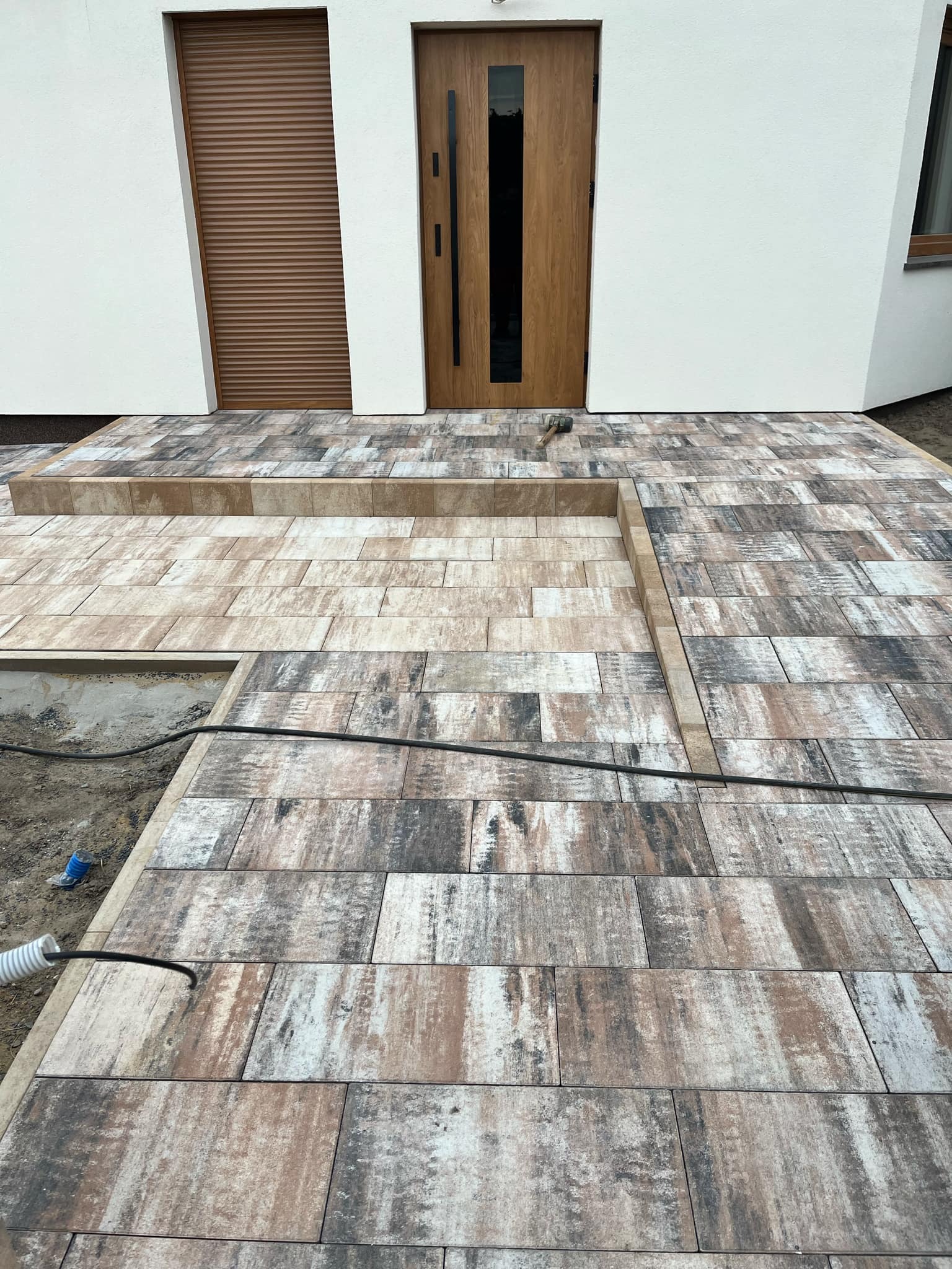 Nowo ułożona kostka brukowa w odcieniach brązu i szarości przed nowoczesnym domem z drewnianymi drzwiami i roletami, widoczny fragment nieukończonego fragmentu z wystającymi kablami.