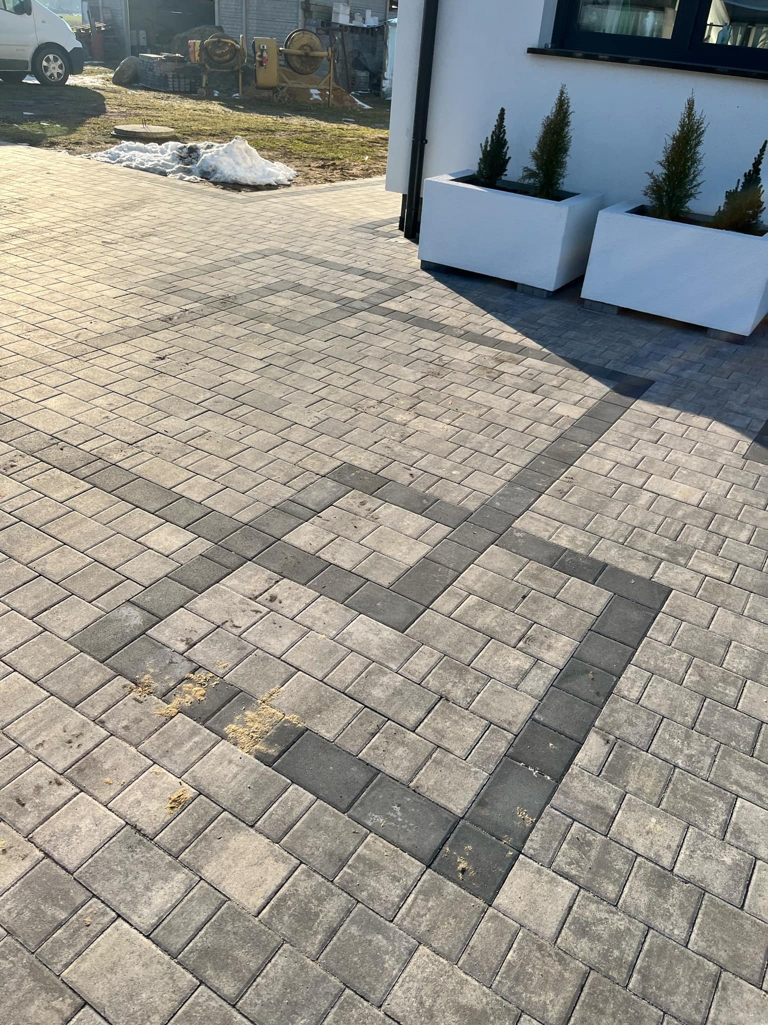 Ułożona kostka brukowa w szarościach z ciemniejszym wzorem, widoczne donice z iglakami i resztki piasku na powierzchni.