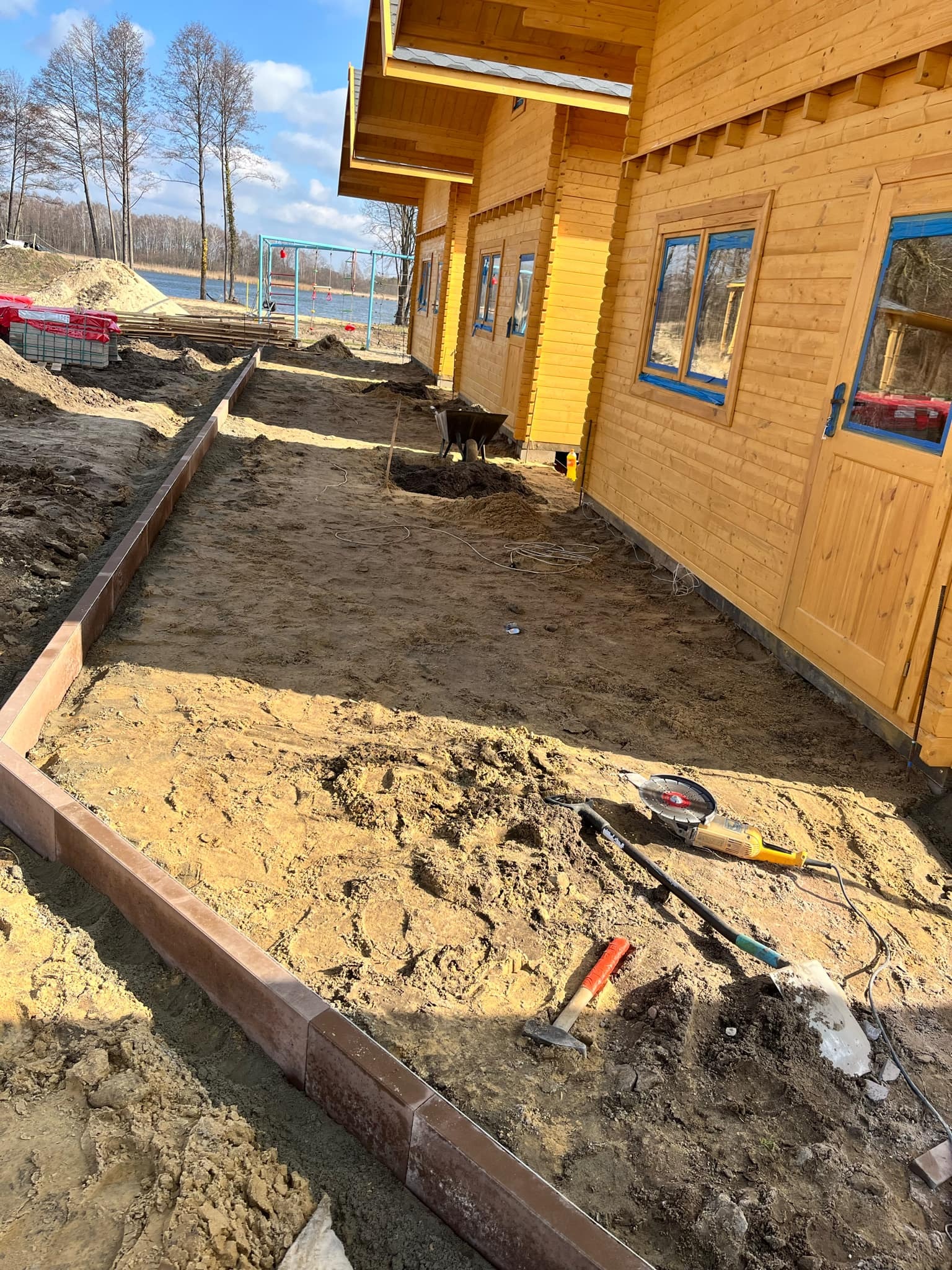 Przygotowanie podłoża pod ścieżkę z obrzeżami wzdłuż drewnianego domu, widoczne narzędzia: szlifierka kątowa, młotek, łopata. W tle jezioro i plac zabaw.