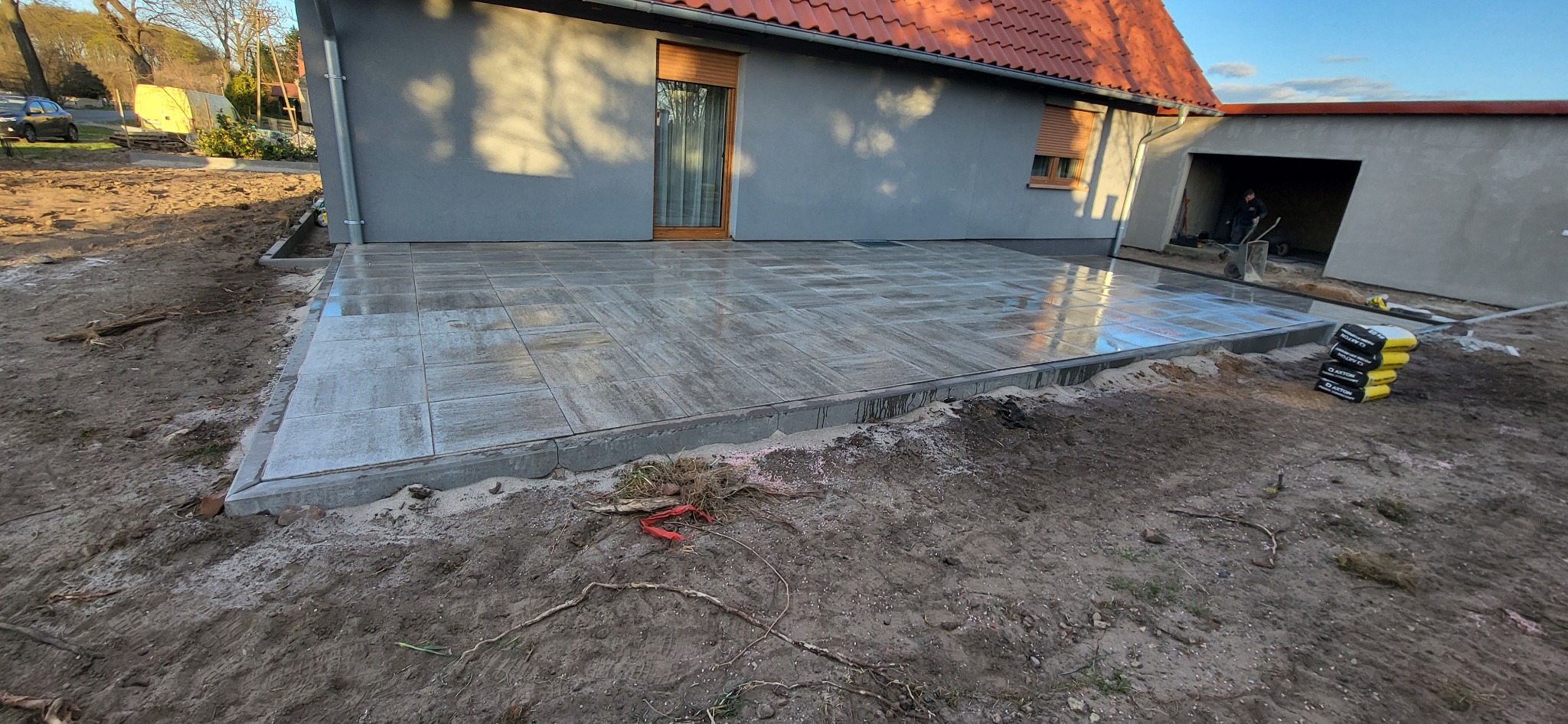 Świeżo ułożony taras z szarych płyt betonowych przy szarym domu z czerwoną dachówką, widoczne worki z zaprawą i pracownik z taczką w tle, teren wokół w trakcie prac ziemnych.
