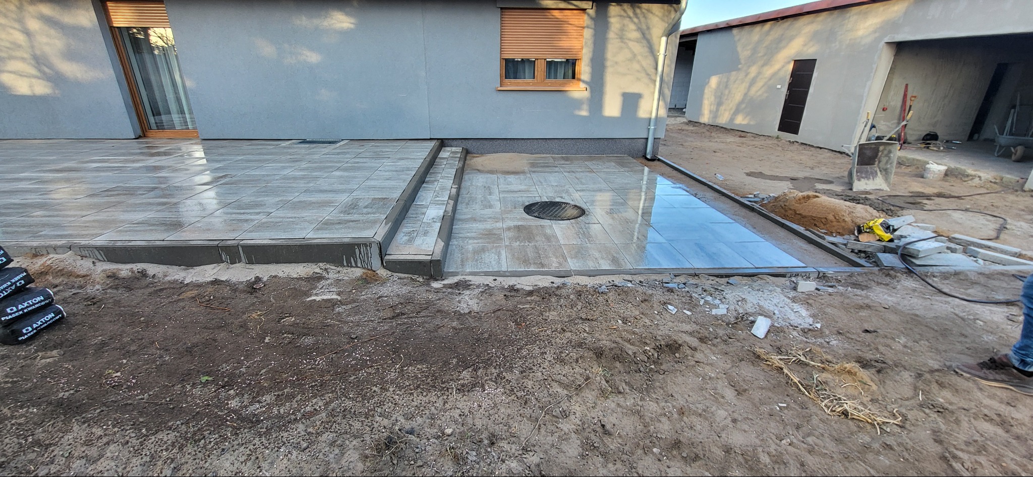 Układanie szarej kostki brukowej na tarasie domu jednorodzinnego, widoczne etapy prac ziemnych i drenaż studzienki kanalizacyjnej.