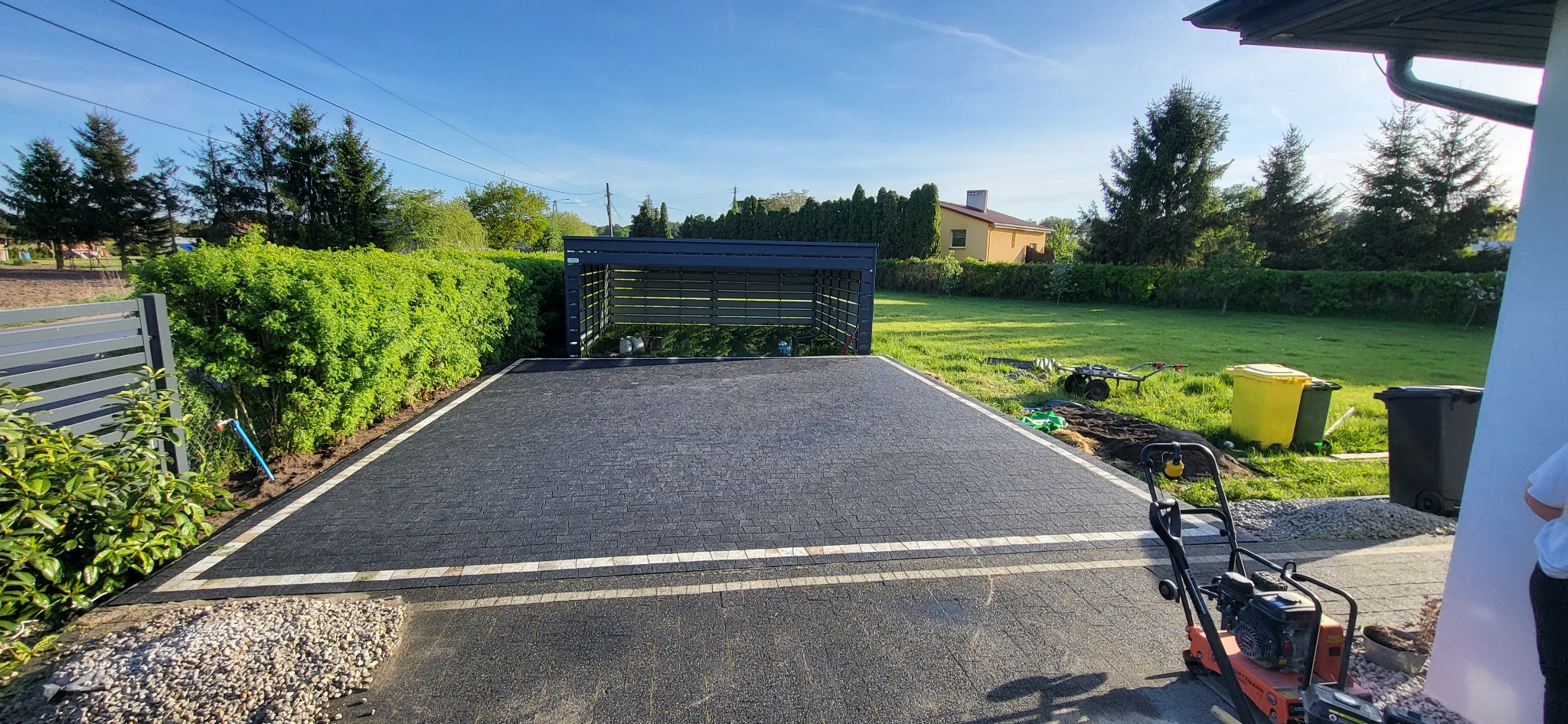 Nowo ułożona, czarna kostka brukowa z jasnymi obrzeżami na podjeździe. W tle widoczny garaż, zieleń i dom. Na pierwszym planie zagęszczarka.