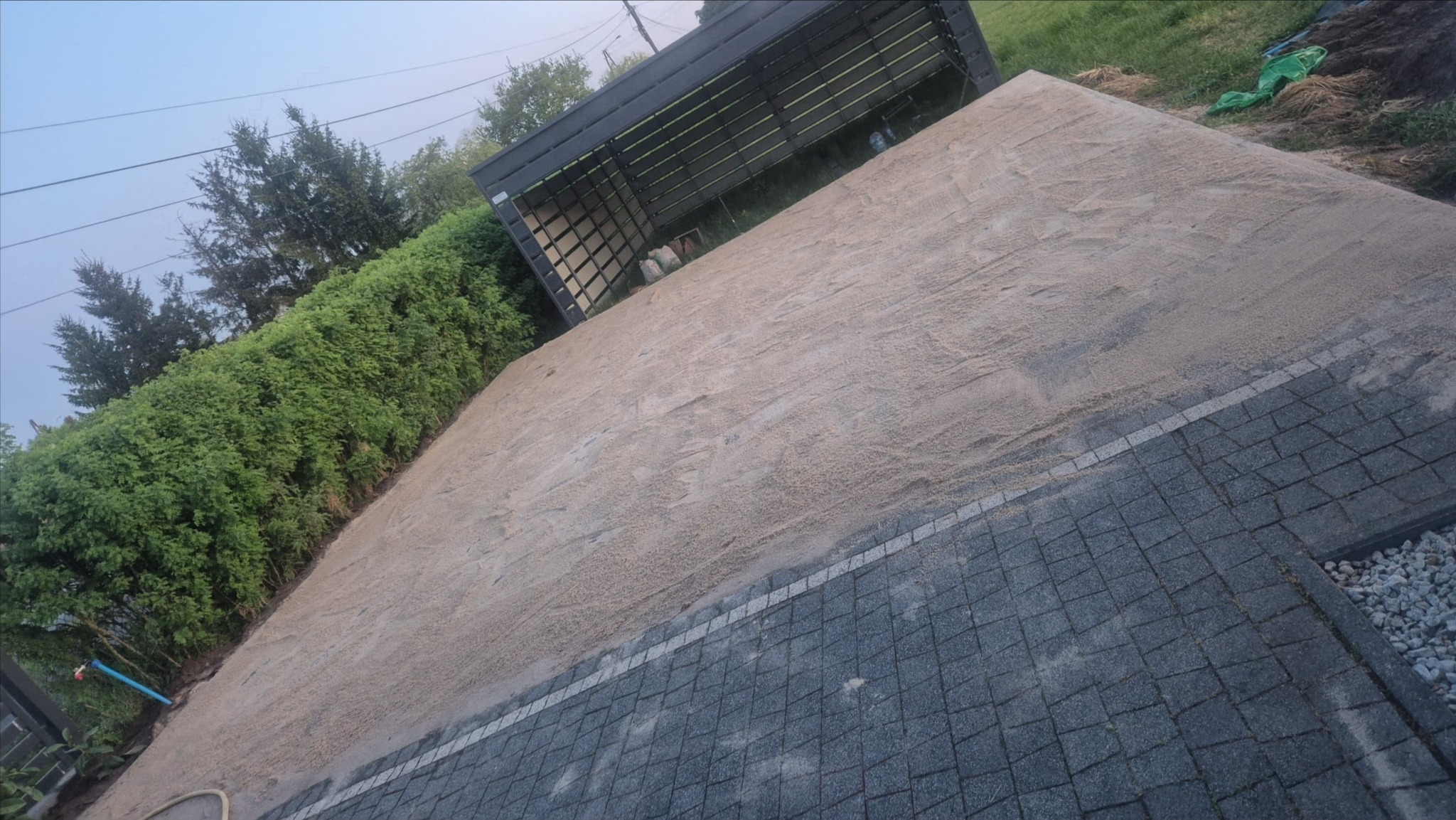 Przygotowany teren pod kostkę brukową z częściowo ułożonym obrzeżem i kostką. Widoczna podsypka piaskowa, pergola i zieleń w tle. Ujęcie z góry.