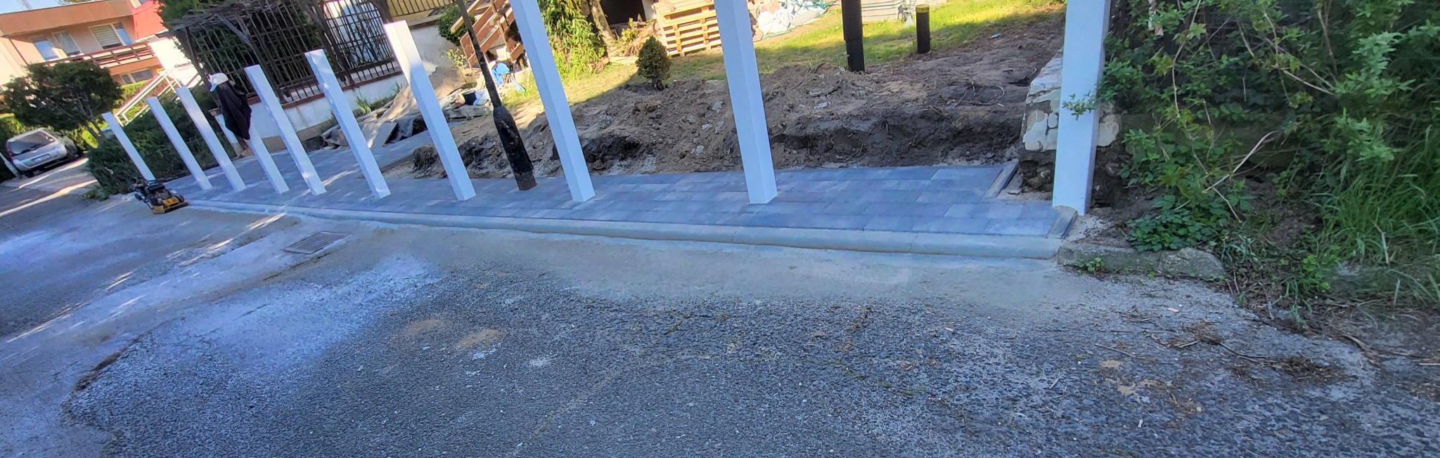 Fragment ułożonej, szarej kostki brukowej przy białych słupkach. W tle widoczna ubijarka wibracyjna i częściowo osoba. Etap prac brukarskich w słoneczny dzień.