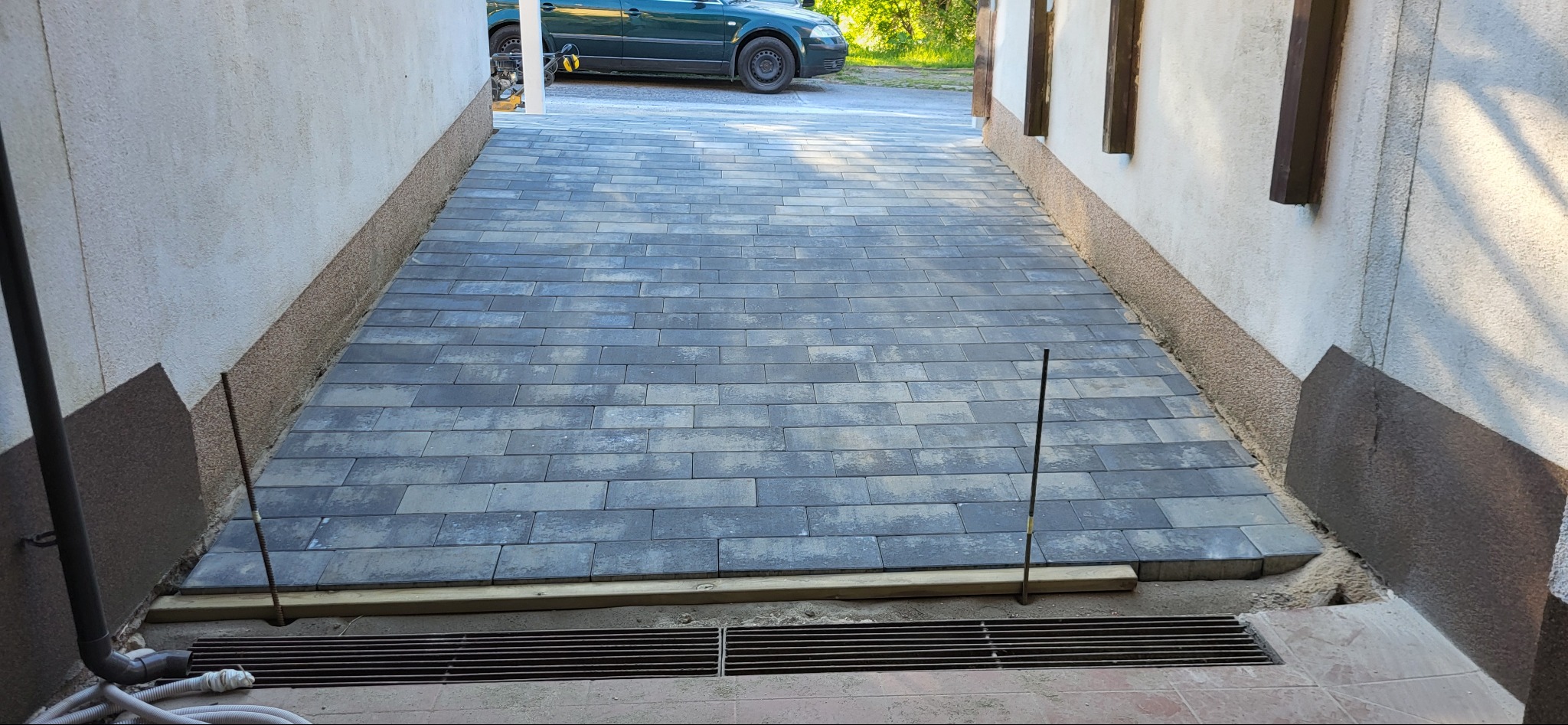 Świeżo ułożona, szara kostka brukowa w wąskim przejeździe między budynkami, z widocznym odpływem liniowym i wystającymi prętami stabilizującymi.
