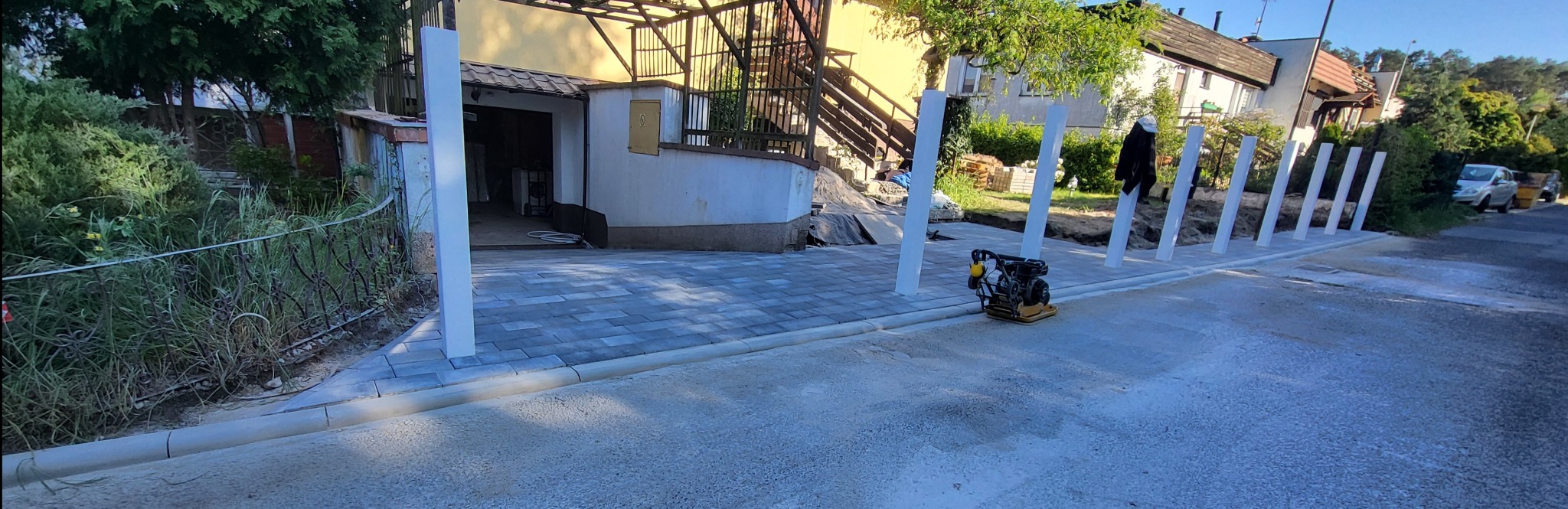 Układanie kostki brukowej na posesji. Widoczna ubijarka, białe słupki graniczne i częściowo ułożona kostka brukowa w szarym odcieniu. Prace w toku.