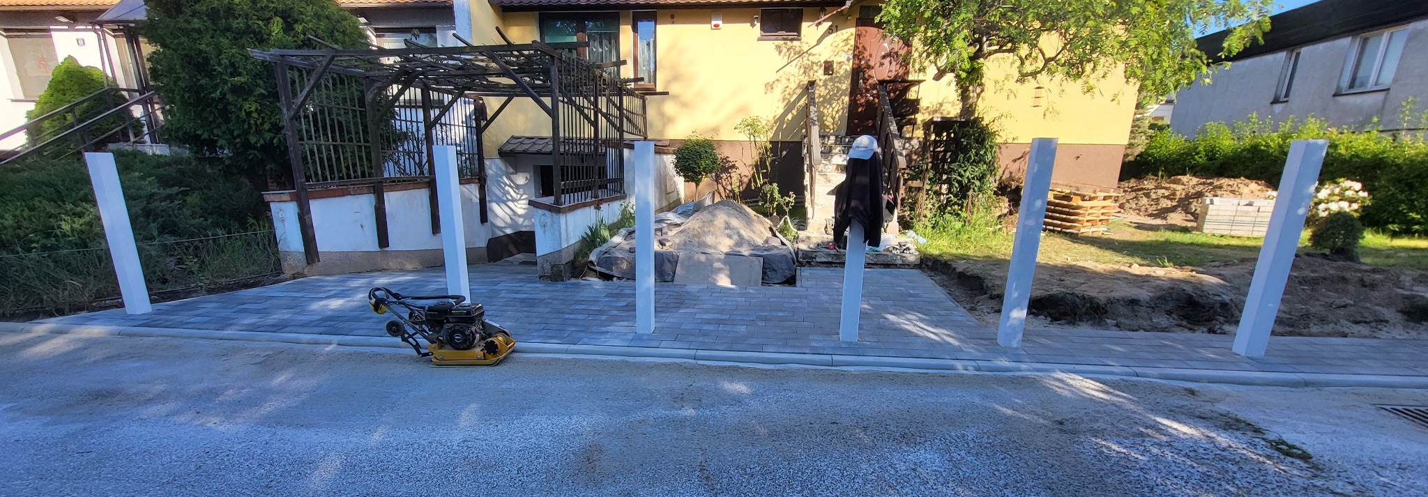 Częściowo ułożona szara kostka brukowa z żółtą zagęszczarką i białymi słupkami w ogrodzie przed domem. Widoczny piasek i ziemia, etap prac.