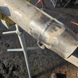 Spaw-Bud Jarosław Komorowski - Szlifowany spaw na metalowej rurze, podtrzymywanej przez statyw spawalniczy, widoczne ślady obróbki i niedoskonałości powierzchni.