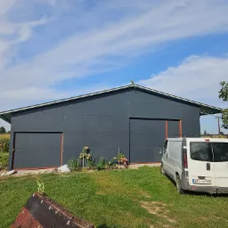 Szara hala z przesuwnymi drzwiami, widok z zewnątrz, obok zaparkowany biały bus, w tle pole kukurydzy.