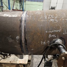 Spaw-Bud Jarosław Komorowski - Spoina na rurze przemysłowej z widocznymi oznaczeniami i elementami mocującymi, umieszczona na palecie w hali produkcyjnej.