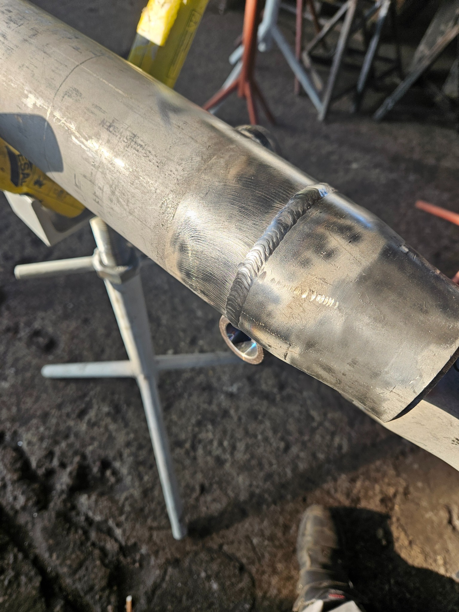 Szlifowany spaw na metalowej rurze, podtrzymywanej przez statyw spawalniczy, widoczne ślady obróbki i niedoskonałości powierzchni.