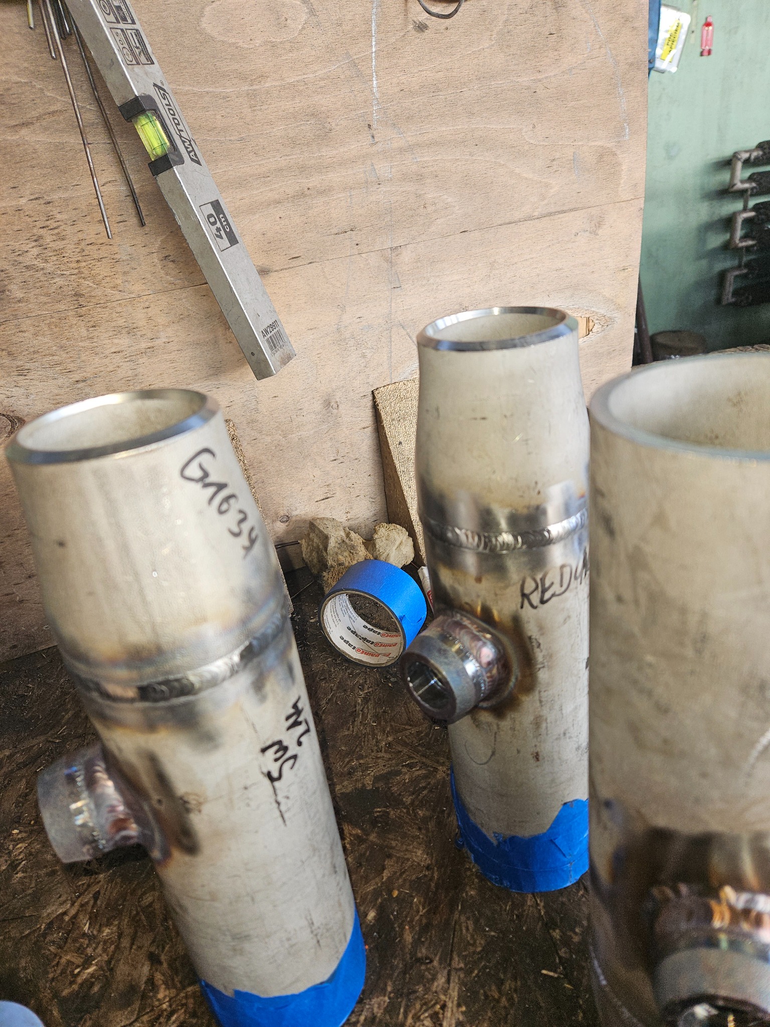 Przygotowane do montażu, pospawane rury ze stali nierdzewnej z oznaczeniami pisakiem, zabezpieczone niebieską taśmą malarską, leżące na warsztatowym stole, obok poziomica