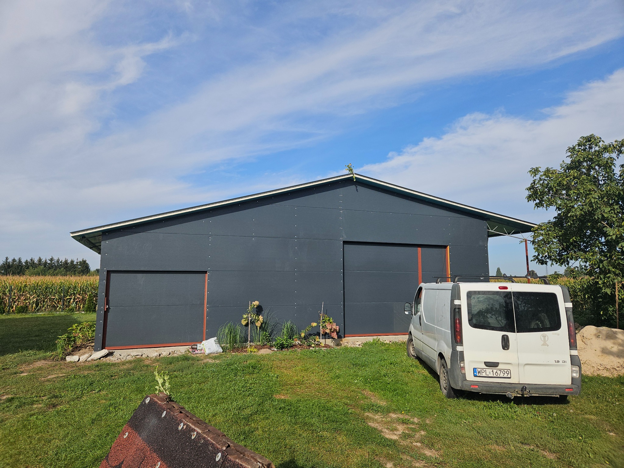 Szara hala z przesuwnymi drzwiami, widok z zewnątrz, obok zaparkowany biały bus, w tle pole kukurydzy.