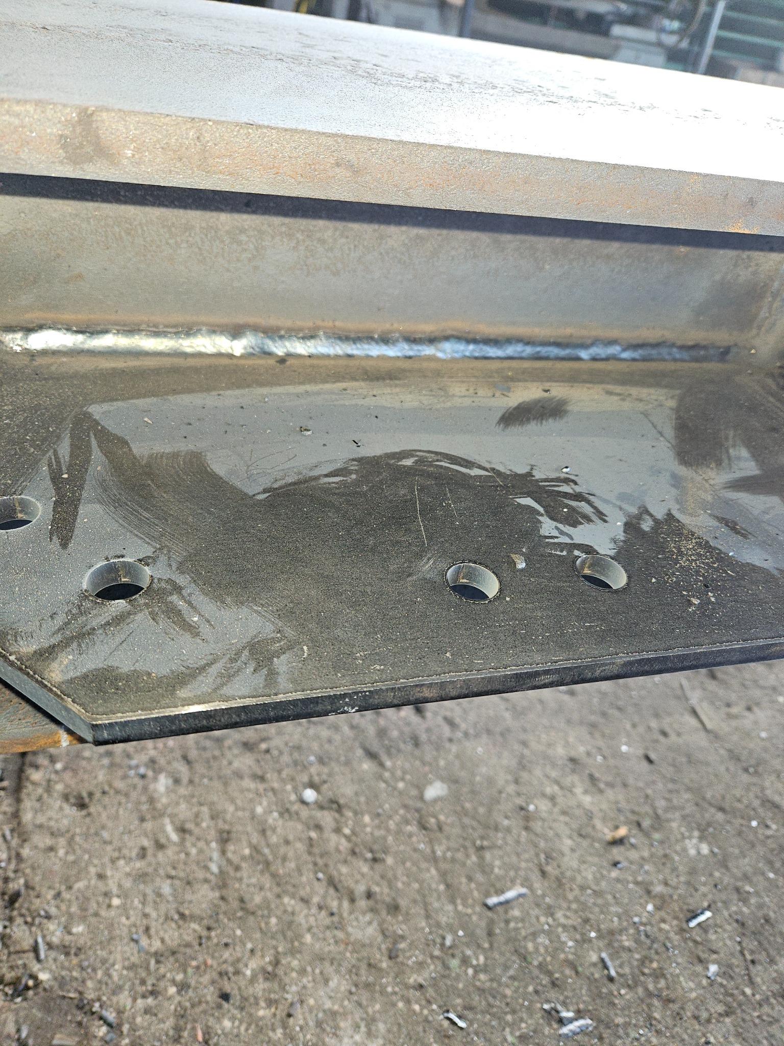 Szczegółowe zbliżenie spawu łączącego dwie stalowe płyty pod kątem prostym, z widocznymi otworami w dolnej płycie, na tle betonowej posadzki.