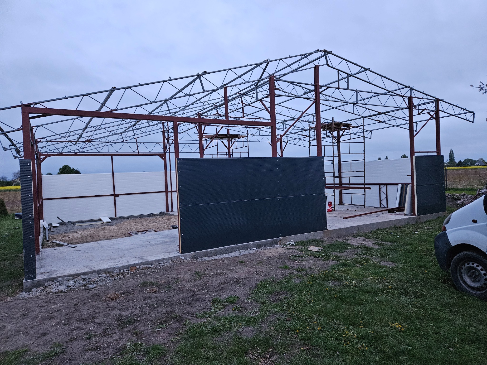 Konstrukcja stalowa budynku w trakcie montażu, widoczne elementy szkieletu, częściowo zamontowane panele ścienne i betonowa podstawa, na zewnątrz trawa i pojazd.