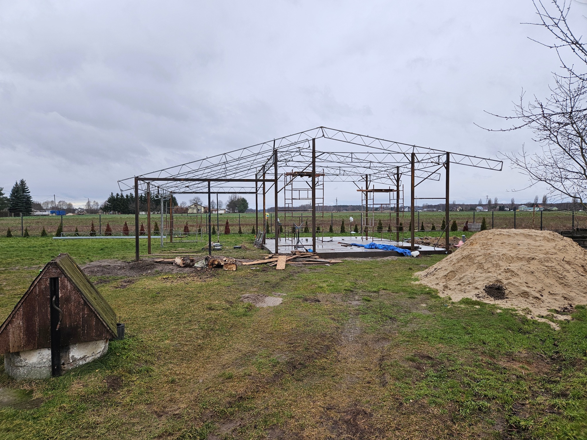 Konstrukcja stalowa szkieletu budynku w trakcie budowy na zewnątrz, widoczny fundament, sterta piasku i studnia zadaszona drewnem na trawiastym terenie w pochmurny dzień.