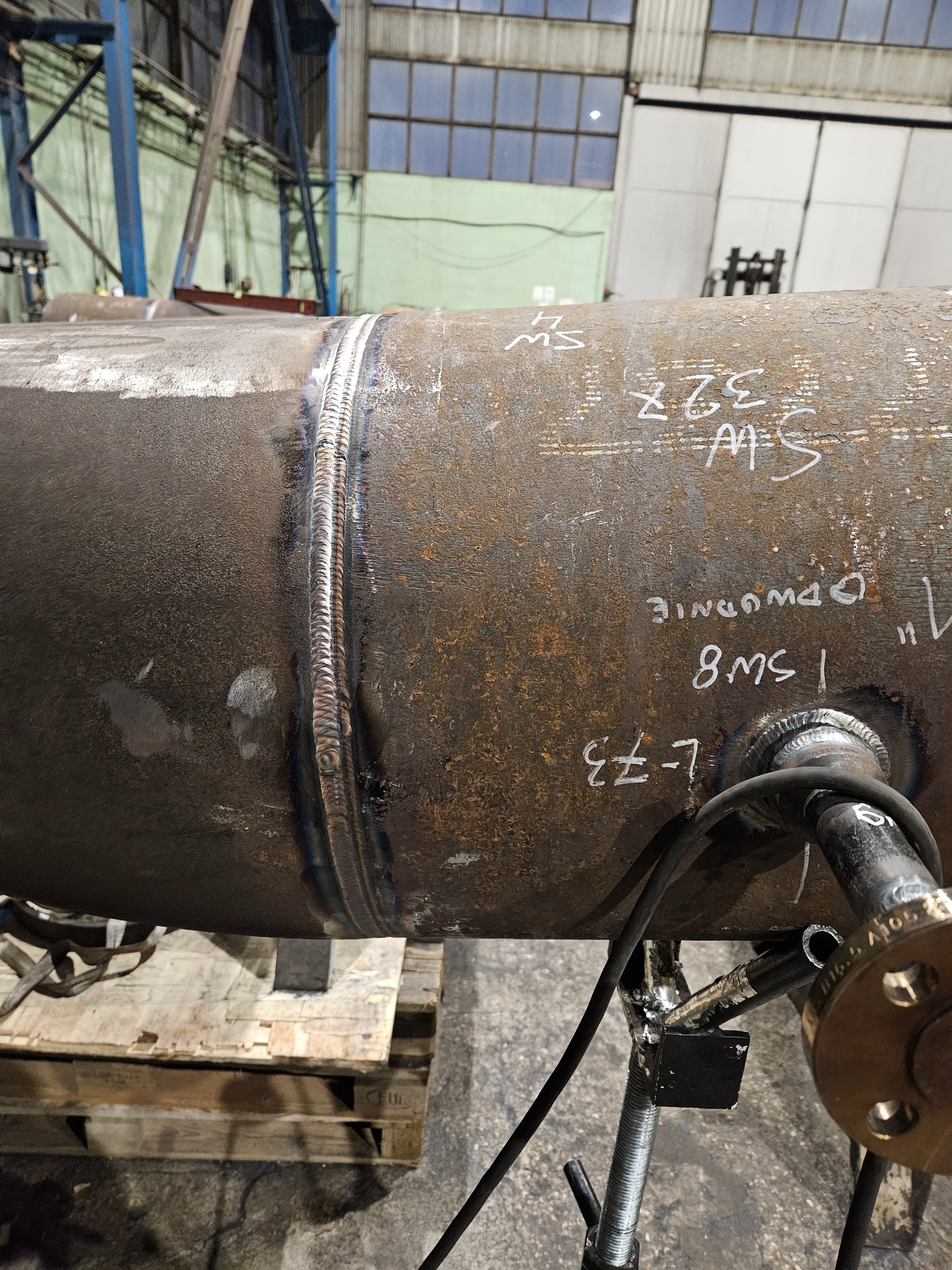 Spoina na rurze przemysłowej z widocznymi oznaczeniami i elementami mocującymi, umieszczona na palecie w hali produkcyjnej.