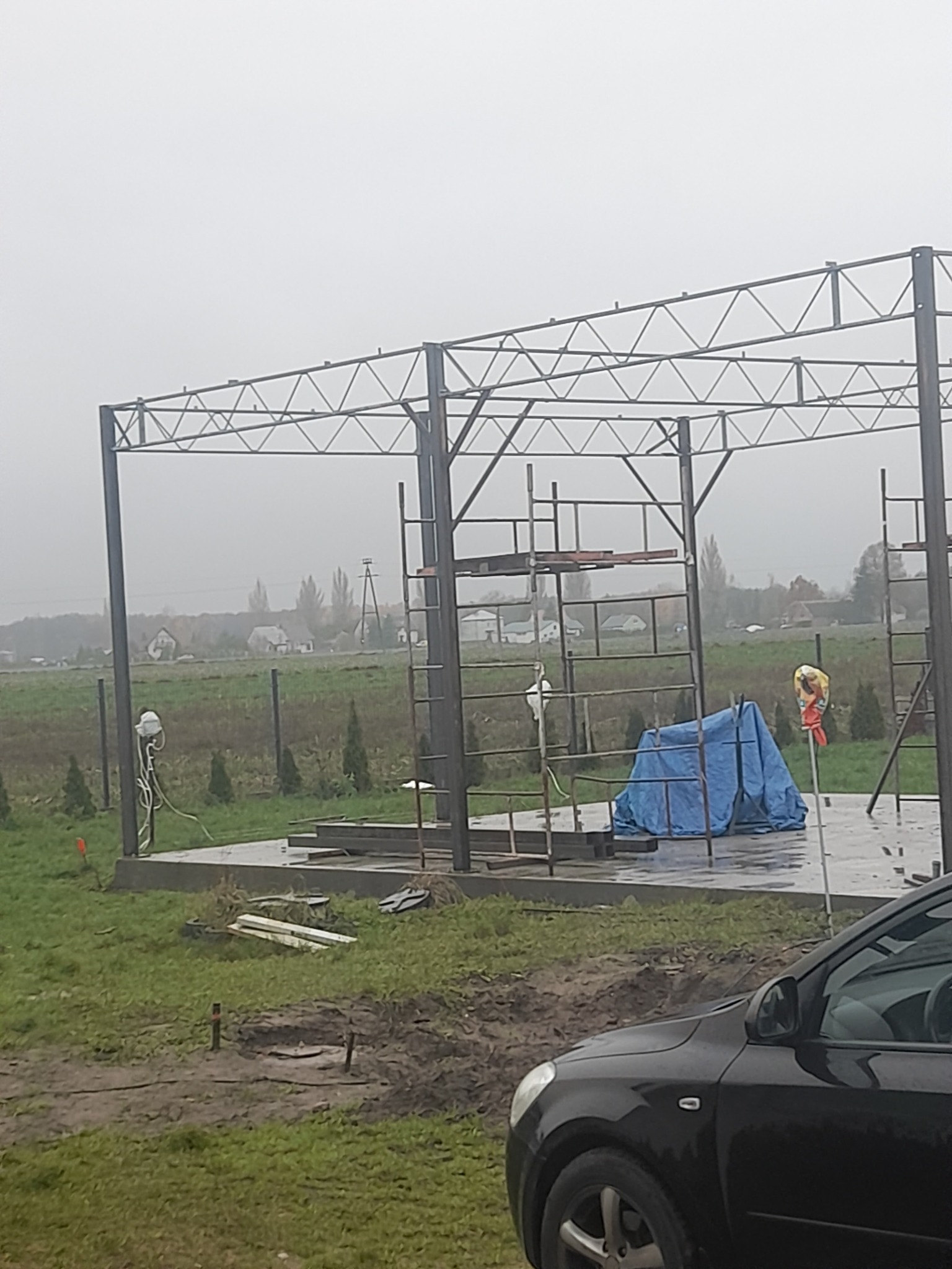 Metalowa konstrukcja zadaszenia w trakcie montażu na betonowej wylewce, widoczne rusztowania i narzędzia, w tle krajobraz wiejski w pochmurny dzień.