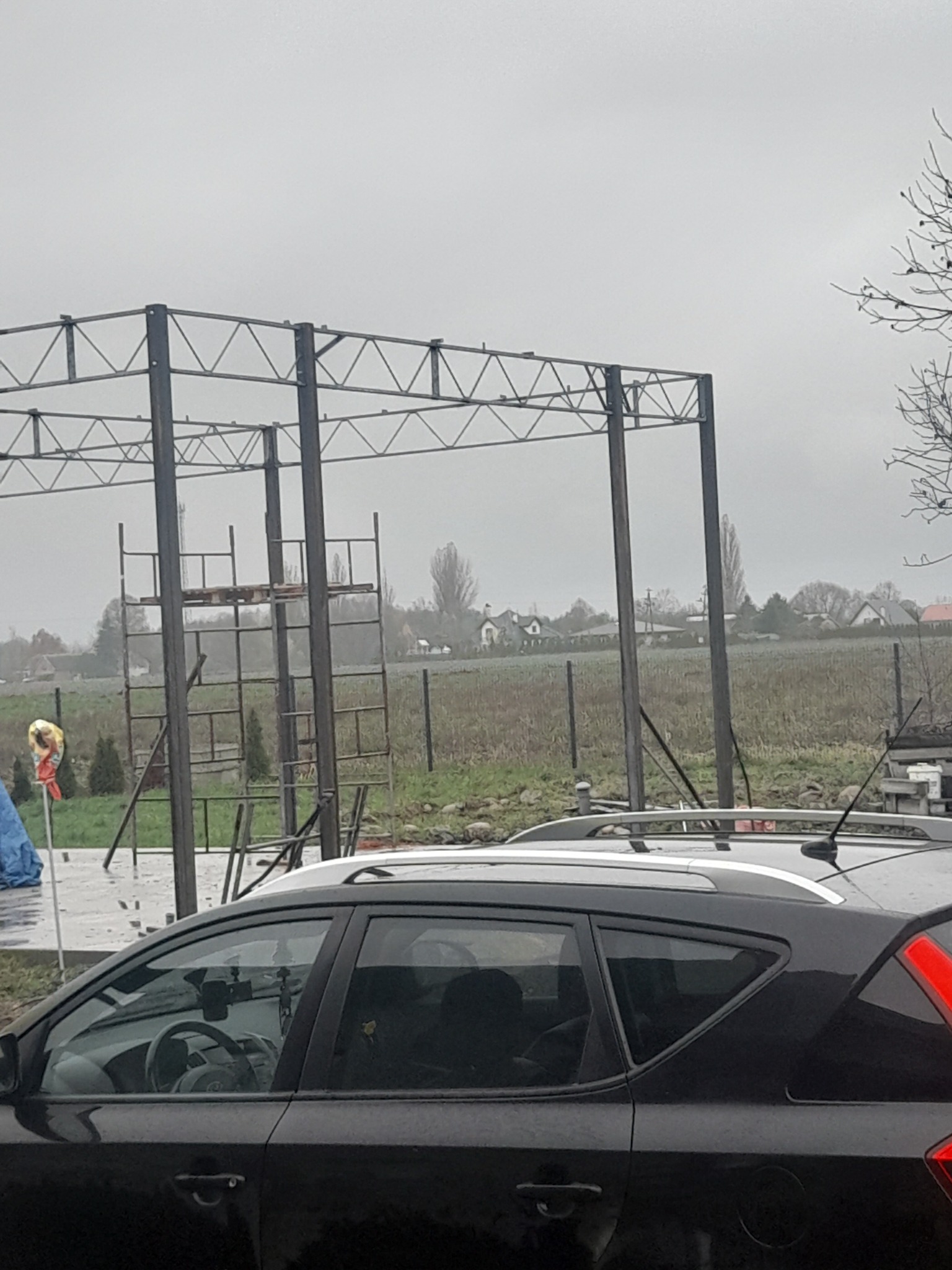 Stalowa konstrukcja z drabinkami w tle, widoczna przez szybę zaparkowanego, czarnego samochodu w pochmurny dzień.