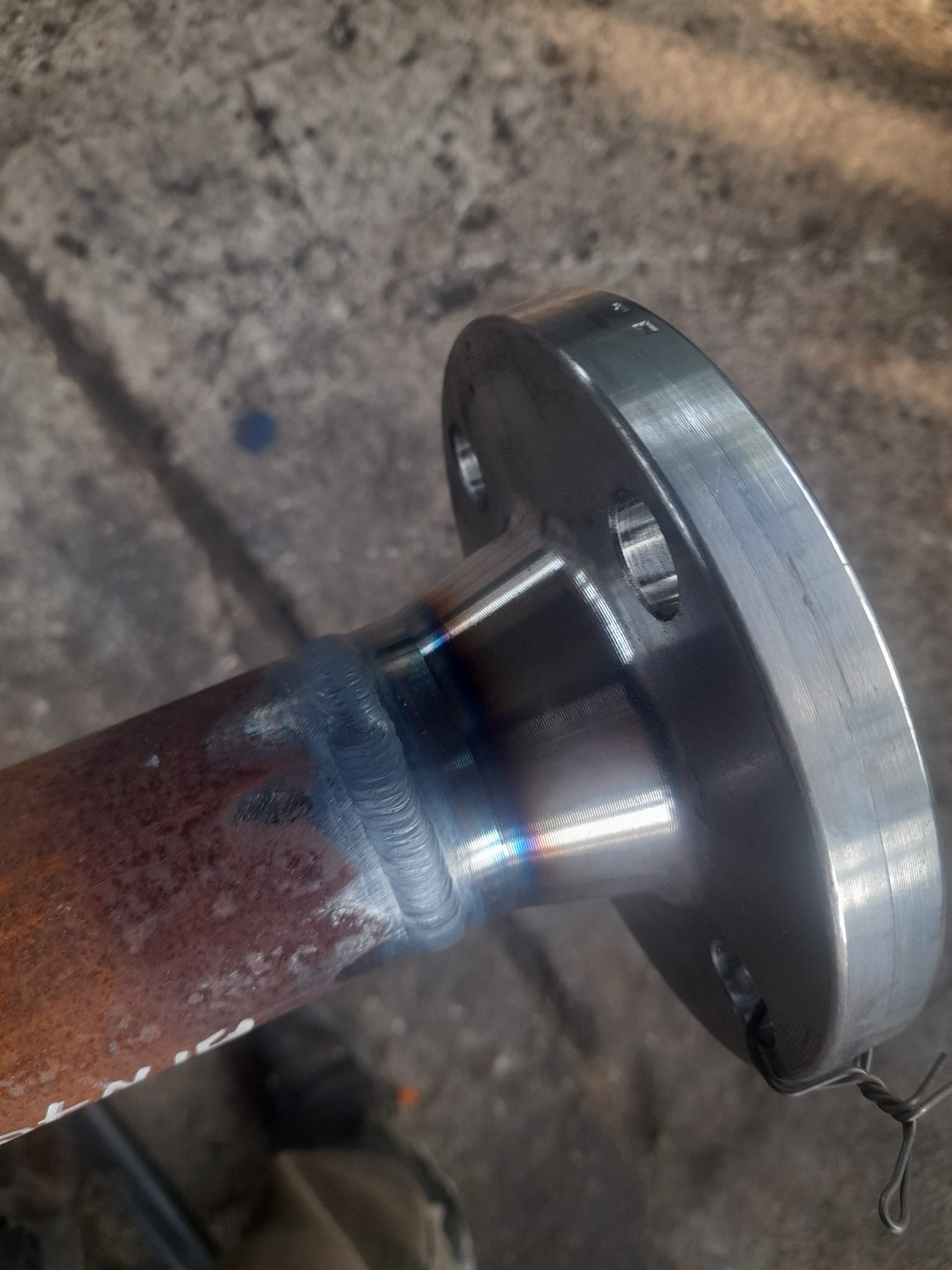 Spoina łącząca rdzewiejącą rurę z metalowym kołnierzem z otworami na śruby, widoczne ślady obróbki i przebarwienia termiczne