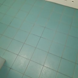 M-J CLEAN - Turkusowe płytki podłogowe w łazience, widoczna fuga i fragment białej szafki z otworami wentylacyjnymi u góry.