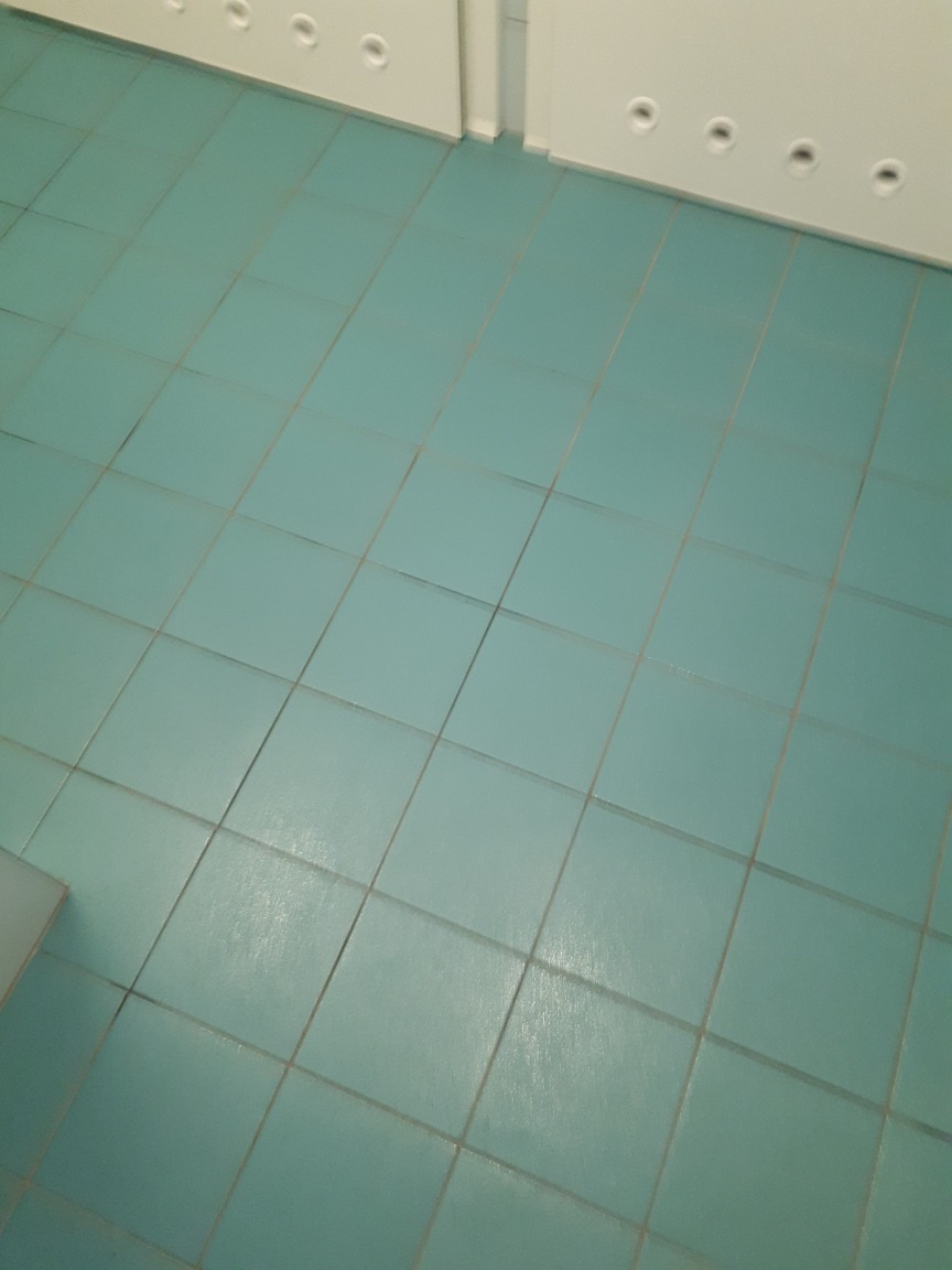 Turkusowe płytki podłogowe w łazience, widoczna fuga i fragment białej szafki z otworami wentylacyjnymi u góry.