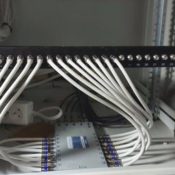 TechnoFix - Wnętrze szafy rack z uporządkowanymi białymi kablami koncentrycznymi podłączonymi do czarnego panelu z numeracją 1-24 i urządzenia rozdzielającego sygnał. Widoczna listwa zasilająca.