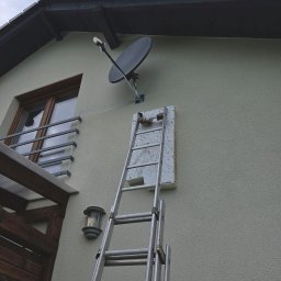 TechnoFix - Czarna antena satelitarna z białym konwerterem zamontowana na beżowej elewacji domu, obok okna z balustradą, z opartą aluminiową drabiną.