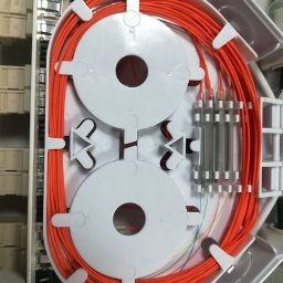 TechnoFix - Wnętrze skrzynki telekomunikacyjnej z pomarańczowymi kablami światłowodowymi, starannie ułożonymi na białych organizerach, widoczne przezroczyste osłonki spawów i terminale.