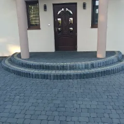 Ułożona kostka brukowa w kolorze grafitowym tworząca półokrągłe schody wejściowe do domu z kolumnami, widoczne okna z roletami i brązowe drzwi wejściowe.