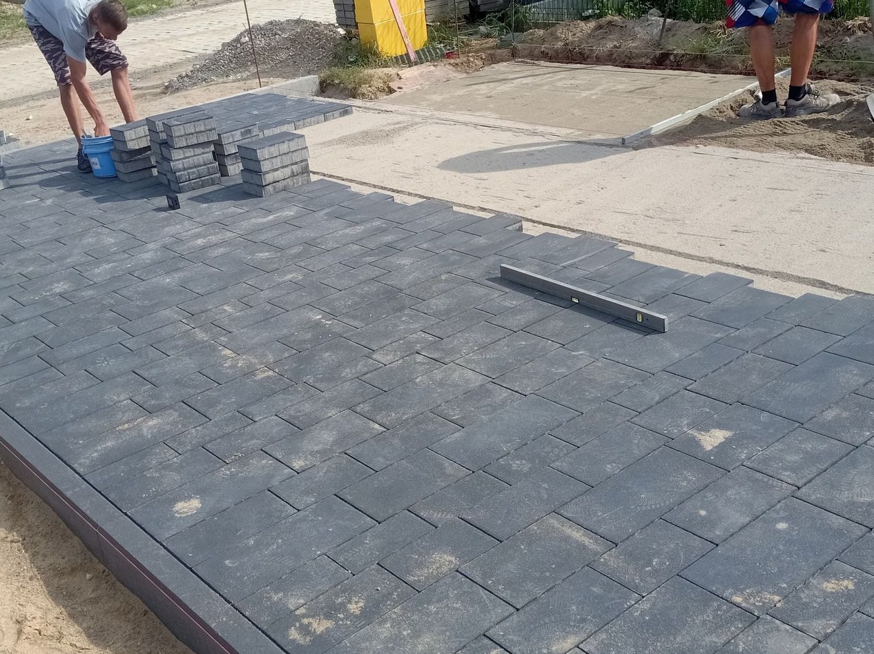 Ciemnoszare prostokątne płyty chodnikowe układane w jodełkę, widoczny pracownik z wiadrem i poziomica, fragment przygotowanego podłoża.