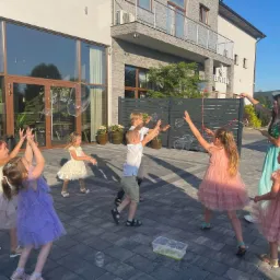 Grupa dzieci w sukienkach bawi się bańkami mydlanymi na zewnątrz, animator w zielonej sukience puszcza bańki za pomocą sznurków, w tle budynek Euforia Dom Weselny.
