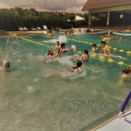 Grupa dzieci i dorosłych bawi się w basenie zewnętrznym z siatką do siatkówki wodnej i fontanną, otoczonym zielenią i budynkiem z zadaszeniem.