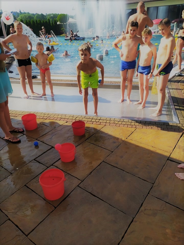 Grupa dzieci w strojach kąpielowych uczestniczy w zabawie z rzucaniem piłeczek do czerwonych wiader ustawionych na mokrym placu, w tle basen z fontanną i innymi osobami.