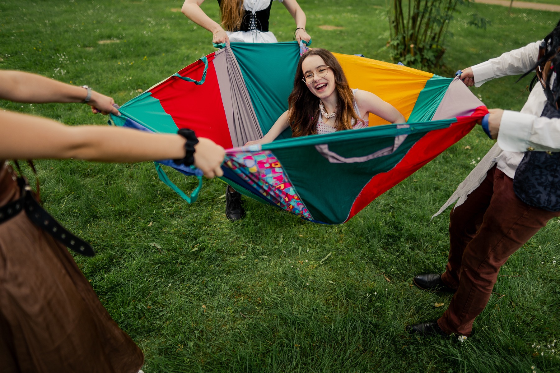Uśmiechnięta dziewczyna w okularach leży w kolorowej chuście trzymanej przez cztery osoby w przebraniach na zielonej trawie podczas animacji dla dzieci.